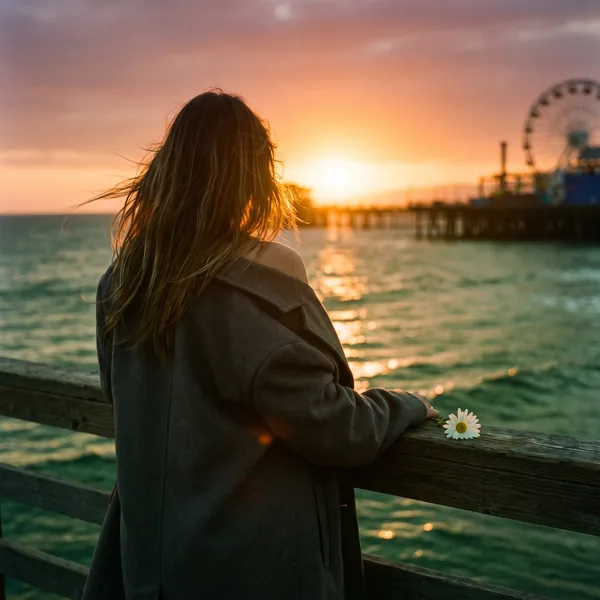 One of One cover art: a woman at the Santa Monica Pier rail at sunset, a white daisy beside her hand